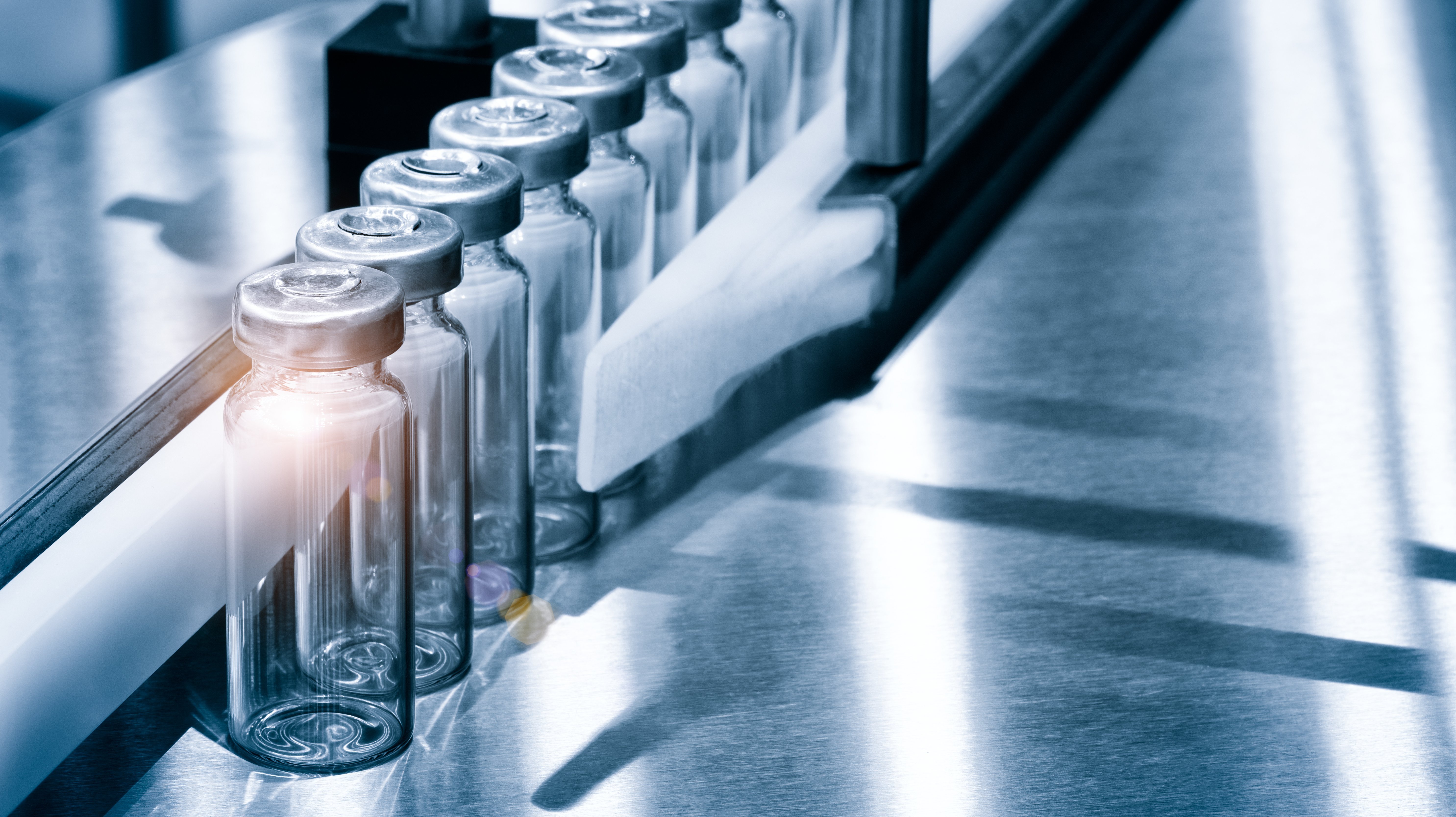 Numerous empy vials lined up in a production machine. The first vial is illuminated by a small glimmer of light.