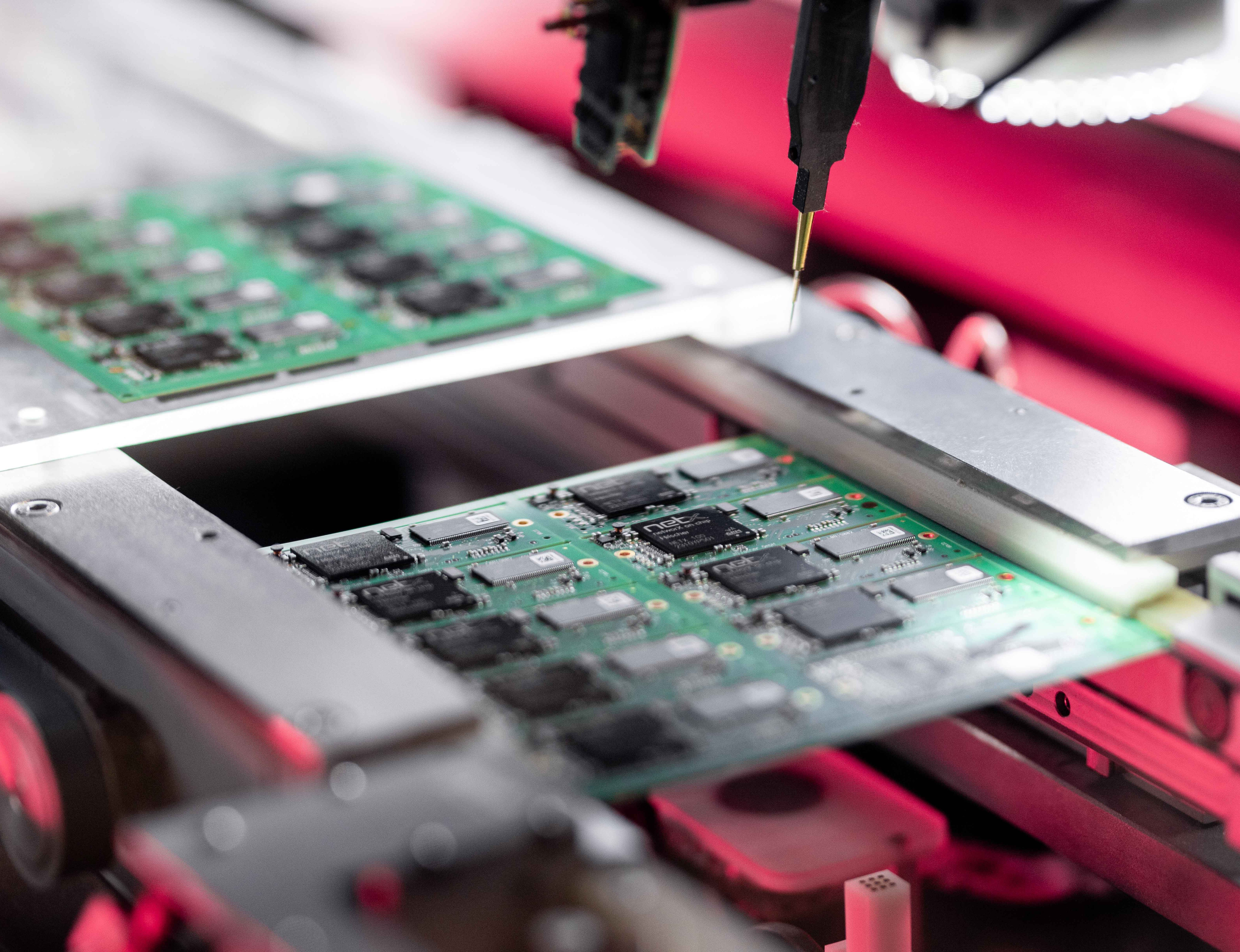 A tray of embedded modules with a netX chip onboard in a production machine. A red gleam is seen in the background. A small golden needle for testing comes from the top pointing at the tray.