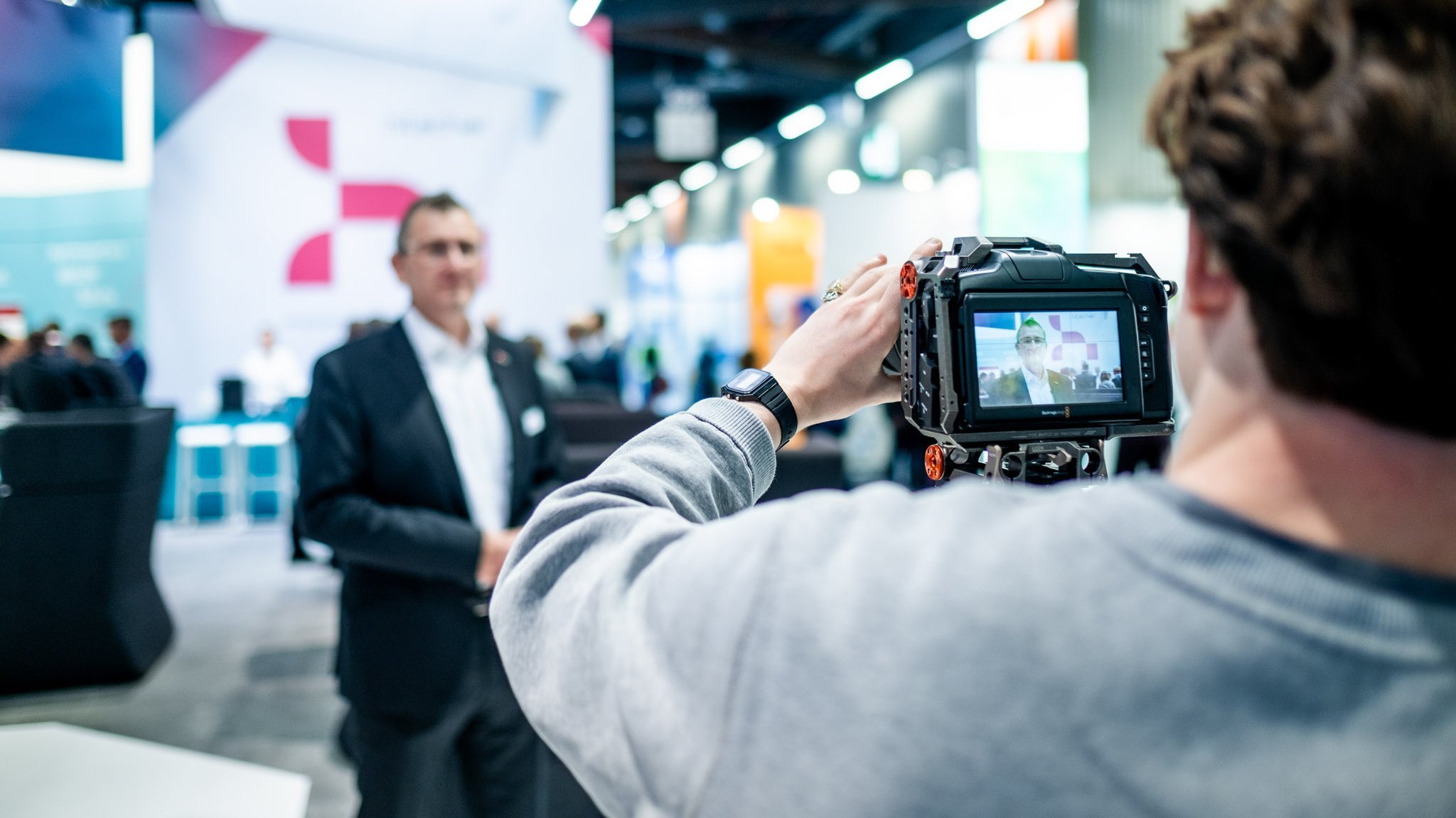 A man from behind operating a camera. He is focussing on a man in a suit a few meters away. In the background is a large Hilscher logo. The photo is taken on a large trade fair booth.