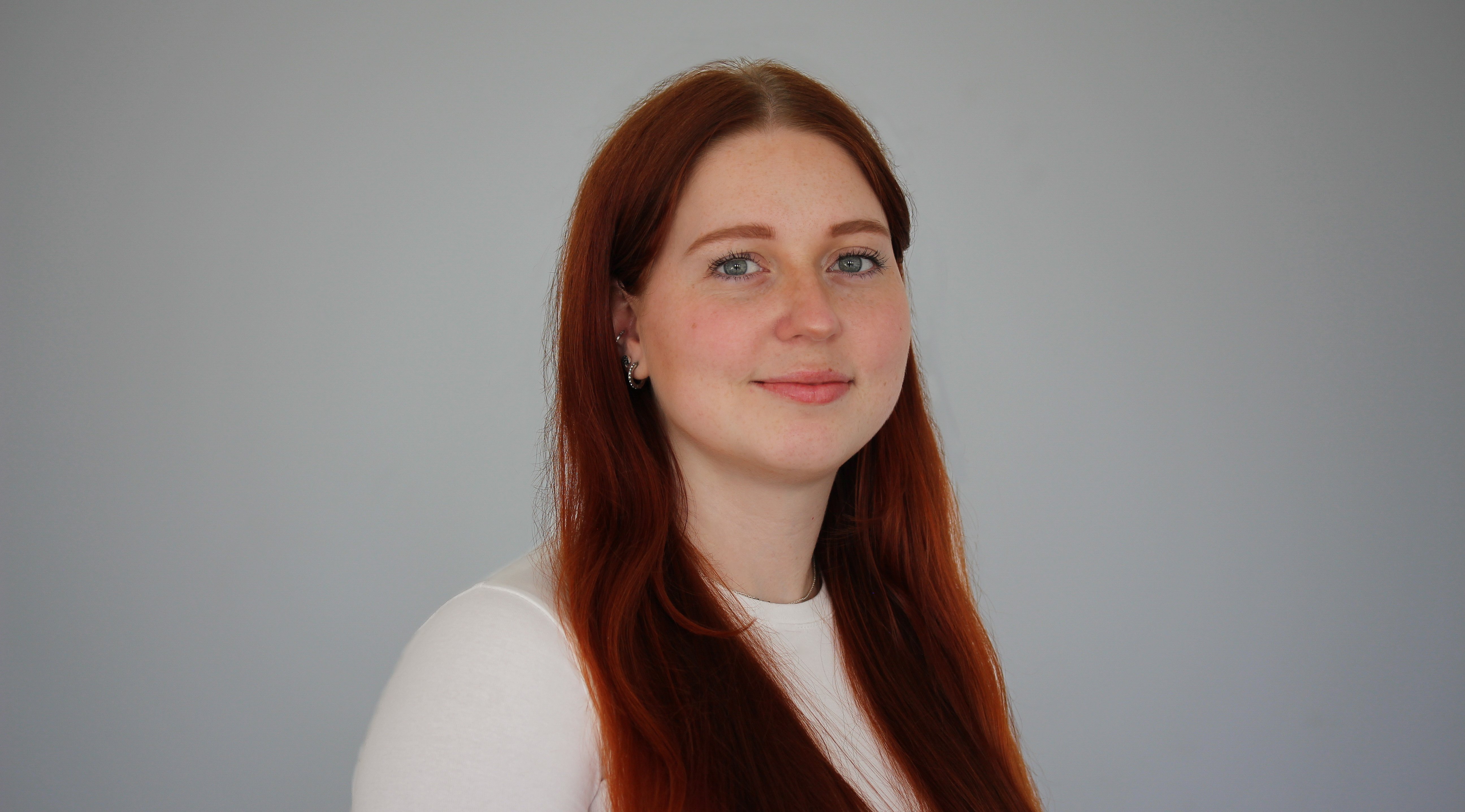 Portrait of a woman with red hair and in a white shirt smiling into the camera. The background is grey.