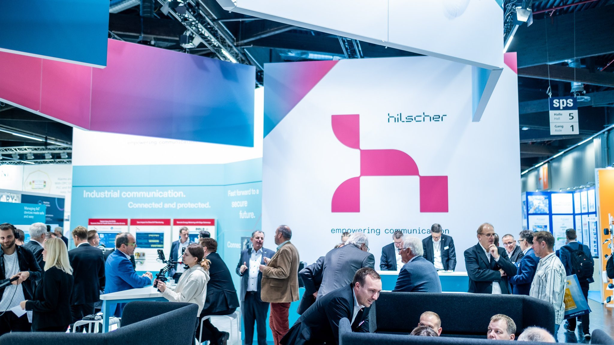 A picture of a crowded trade fair booth. In the background is a large red Hilscher logo on a white wall.