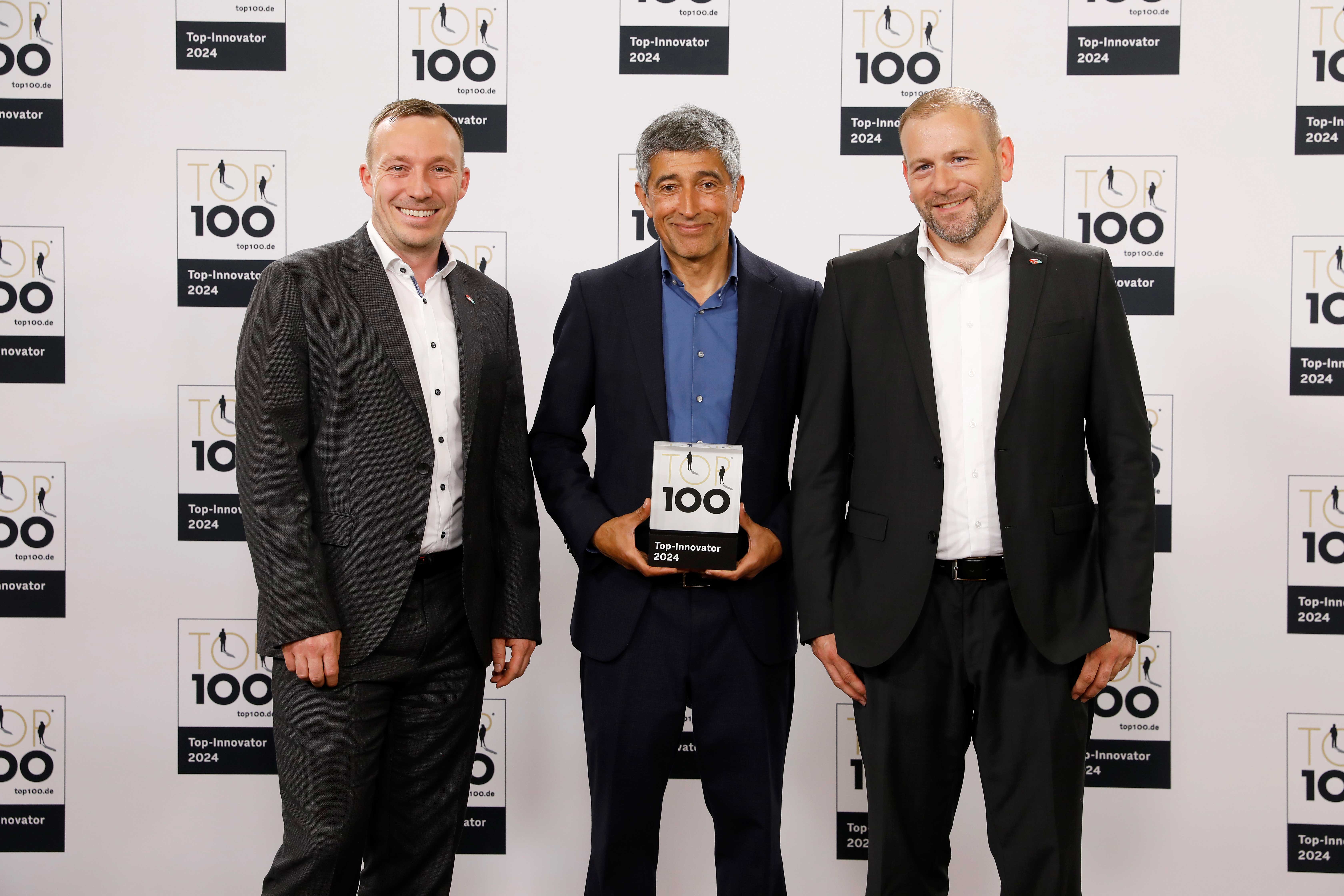 Three men in businesss suits are standing in front of a photo wall. All three are smiling. The man in the middle holds a TOP 100 trophy. In the background are several TOP 100 logos printed on to the wall.