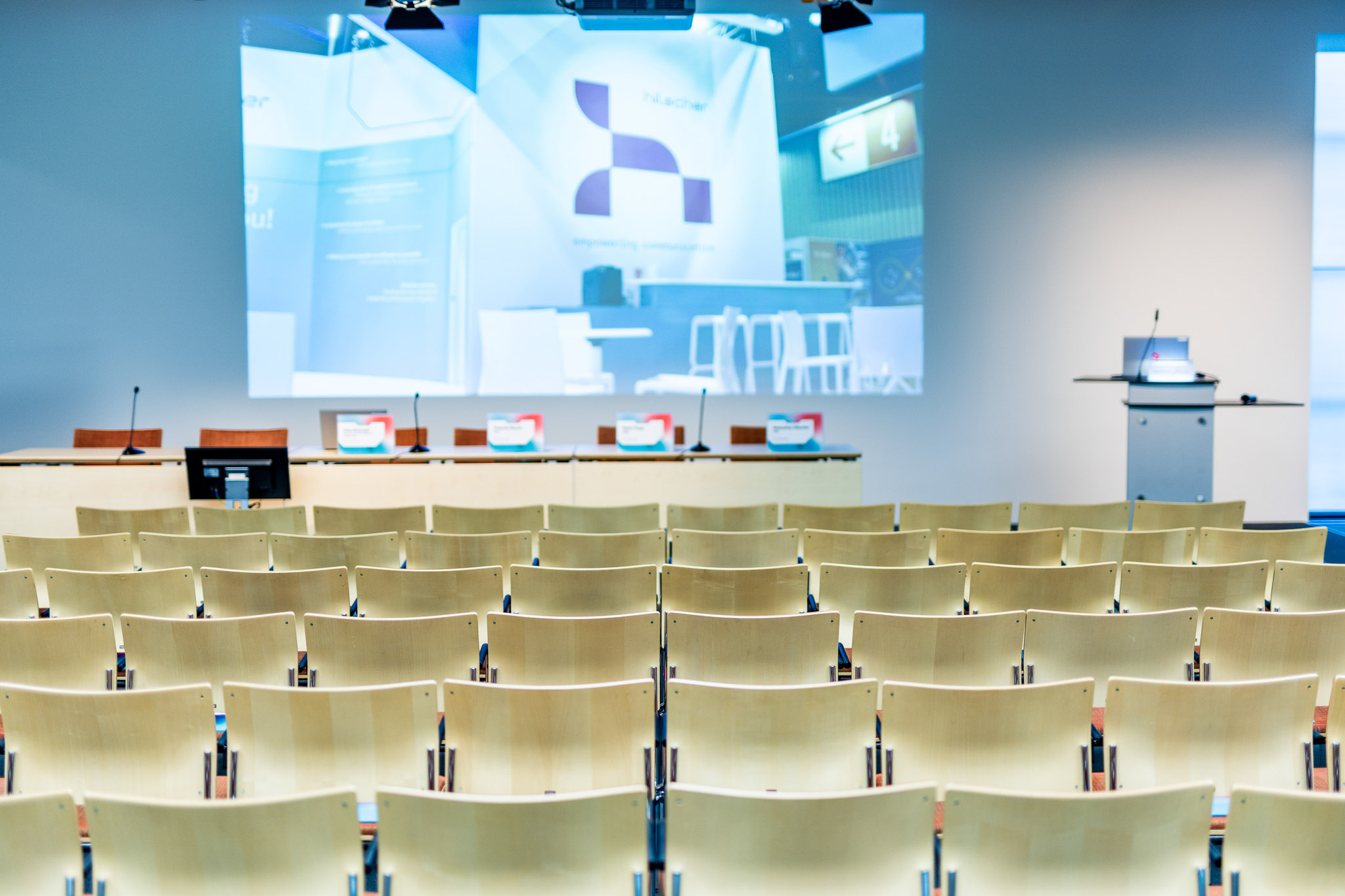 A press conference with many chairs in the foreground. In the background is a stage with several microphones, a desk and a large projection of the Hilscher logo. The room is empty.